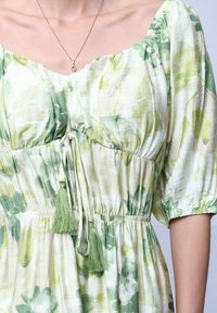 Robe fleurie en vert clair et blanc avec des manches bouffantes courtes, un décolleté à cordon de serrage et des accents en pompons verts. Tissu texturé et léger.