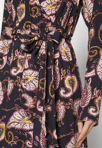 Černé šaty s fialovým, žlutým a bílým paisley vzorem, s uvázaným tkaninovým opaskem v pase a dlouhými rukávy, ruka opřená o bok.