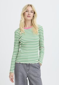 Frau mit langen blonden Haaren, die ein grün-weiß gestreiftes Langarmshirt mit pinkem Kragen und grauen Jeans trägt, steht mit einer Hand in der Tasche.