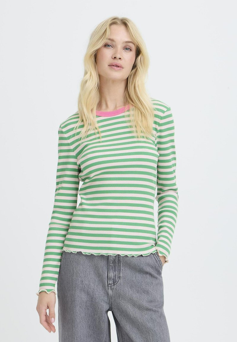 Frau mit langen blonden Haaren, die ein grün-weiß gestreiftes Langarmshirt mit pinkem Kragen und grauen Jeans trägt, steht mit einer Hand in der Tasche.