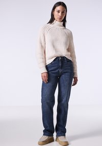 Suéter de punto acanalado color crema con cuello alto, jeans de mezclilla oscuros de cintura alta, combinado con zapatos de lona beige texturizados. Enfocado en un ajuste relajado y comodidad.
