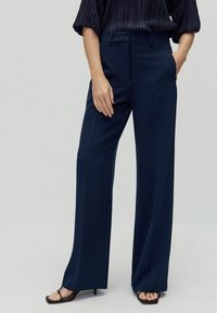 Marineblå brede bukser lavet af glat stof, med høj talje og sidelommer, parret med sorte sandaler med tynde stropper.