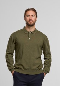 Bearded man with long hair in an olive green long-sleeve polo shirt and dark pants, standing with hands in pockets against a plain background.