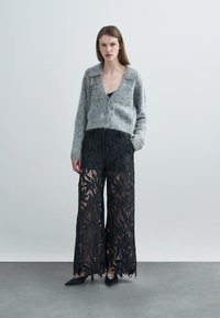 Cardigan gris duveteux avec un décolleté en V profond, associé à un pantalon large noir orné de motifs complexes de feuilles. Chaussures noires à bout pointu.