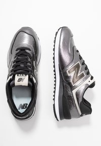 Sneakers New Balance in pelle nera e grigio metallico, con suola bianca, lacci neri e dettagli con logo. Superficie con finitura testurizzata.