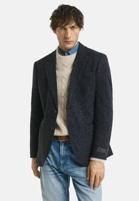 Uomo in giacca scura a macchie, maglione lavorato a trecce crema, camicia di jeans blu e jeans azzurri su sfondo bianco semplice.