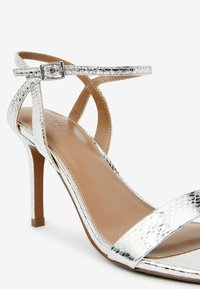 Sandal con tacco alto in argento con una finitura lucida effetto pelle di serpente, cinturino alla caviglia regolabile e un design elegante con punta affusolata.