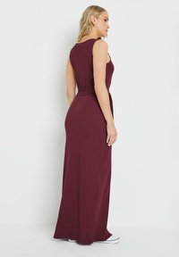 Robe maxi sans manches bordeaux avec un corsage ajusté et une ceinture à nouer, présentant une texture côtelée et une fente sur le côté, associée à des baskets blanches.