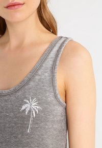 Camiseta sin mangas gris con una textura suave, que presenta un gráfico de una palmera blanca en el frente y amplios agujeros para los brazos para mayor comodidad.