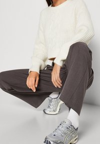 Weißer, kabelgestrickter, cropped Pullover, graue weit geschnittene Hose und silberne Sportschuhe mit Netz- und Gummielementen. Die Hände ruhen auf dem Bein.
