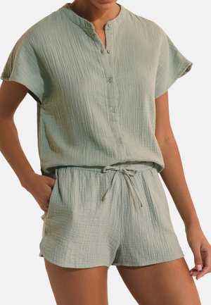 Kobieta w jasnorozowej, teksturowanej koszuli z krótkim rękawem i guzikami oraz pasujących szortach ze ściągaczem, stoi z jedną ręką w kieszeni.