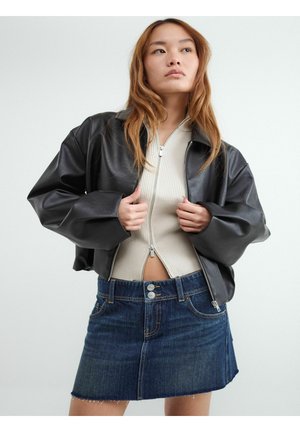 Młoda kobieta poprawia czarną skórzaną kurtkę na beżowej bluzce na suwak i ciemnoniebieskiej dżinsowej spódnicy na jednolitym tle.