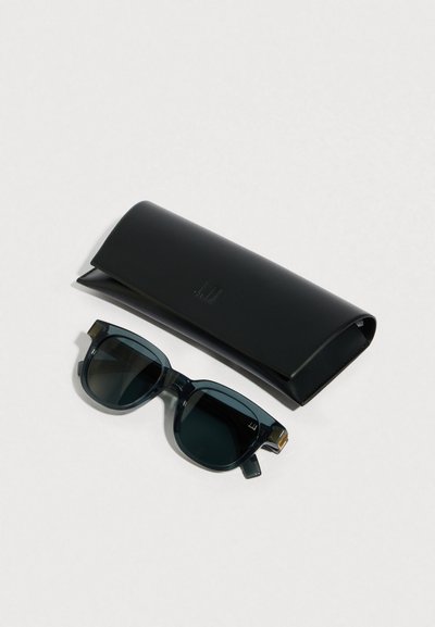 Gafas de sol rectangulares negras con lentes oscuras junto a un estuche de cuero negro plegado sobre una superficie gris clara.