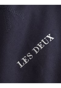 Tissu bleu marine avec broderie "LES DEUX" en texte blanc. Texture lisse avec des détails de couture soignés, mettant en avant un design simple et moderne.
