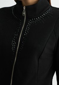 Giacca con cerniera nera caratterizzata da un collo alto, decorata con borchie nere disposte a raggiera sulla parte anteriore. Tessuto dalla texture liscia.