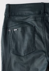 Pantalones de mezclilla verde oscuro con un solo bolsillo trasero que presenta detalles de costura y una pequeña etiqueta metálica. Textura suave y diseño ajustado.