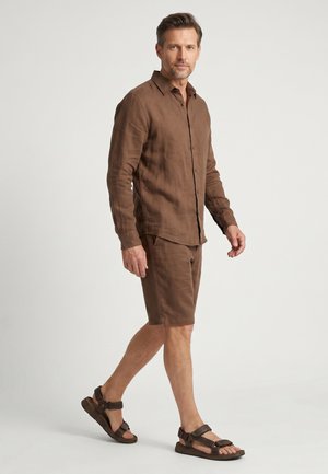 Mann, der geht und ein braunes langärmeliges Hemd mit Knöpfen, passende Shorts sowie braune Sandalen mit offenen Zehen vor einem schlichten hellen Hintergrund trägt.