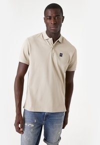 Beige poloshirt met een textuurafwerking en een kraag, met een klein marineblauw logo. Draagt met versleten blauwe jeans.