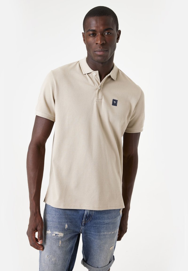 Beige poloshirt met een textuurafwerking en een kraag, met een klein marineblauw logo. Draagt met versleten blauwe jeans.