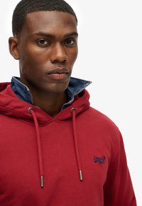 Felpa rossa con cappuccio e coulisse, con un colletto in denim sotto. Logo ricamato sul petto, tessuto di cotone con una superficie liscia.