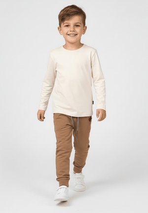 Uśmiechnięty chłopiec idący przed siebie, ubrany w beżową koszulę z długim rękawem, brązowe spodnie dresowe i białe tenisówki na jednolitym tle.