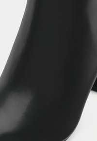 Botín negro hasta el tobillo con un acabado brillante, forma contorneada y textura suave. Presenta un material similar al cuero con un diseño minimalista.