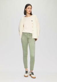 Kardigan z kości słoniowej z guzikami, jasnobeżowa koszula z grafiką serca, zielone wąskie dżinsy oraz kolorowe sneakersy z sznurówkami.