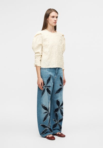 Λευκή κρεμ μπλούζα με υφή και μακριά μανίκια, με συγκεντρωμένους ώμους, σε συνδυασμό με μπλε τζιν παντελόνι φαρδιάς γραμμής με σκοτεινές τζιν floral εφαρμογές. Καφέ παπούτσια.
