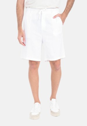 Uomo che indossa pantaloncini bianchi con coulisse e sneakers bianche, in piedi con una mano in tasca contro uno sfondo chiaro e uniforme.