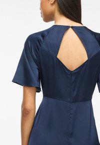 Frau trägt ein dunkelblaues Kleid mit kurzen, flatternden Ärmeln und einem rautenförmigen offenen Rücken, der am Hals geschlossen ist, sowie einen Reißverschluss an der Taille.