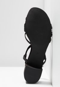 Sandale noire à lanières avec semelle texturée et talon carré, vue de dessous sur fond blanc en coin.