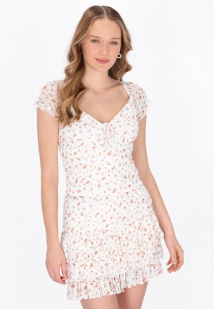 Tânără cu părul lung și ondulat, purtând o rochie albă scurtă cu imprimeu floral, tiv cu volane și mâneci evazate, stând în picioare pe un fundal simplu.