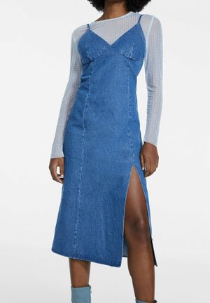 Femme portant une robe midi en denim bleu avec une fente haute sur la cuisse, portée sur un top blanc transparent à manches longues avec petits pois.