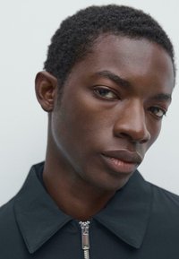 Nærbillede af en ung sort mand med kort krøllet hår, iført en mørk jakke med lynlås foran, der kigger let til siden mod en ensfarvet baggrund.