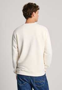 Sudadera de color crema, de mangas largas, con puños y dobladillo acanalados, textura suave. Combinada con jeans de mezclilla azul, mostrando la vista trasera.