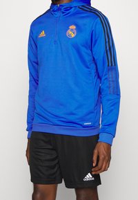 Homme portant un haut d'entraînement bleu Adidas du Real Madrid avec un short noir Adidas sur fond blanc.