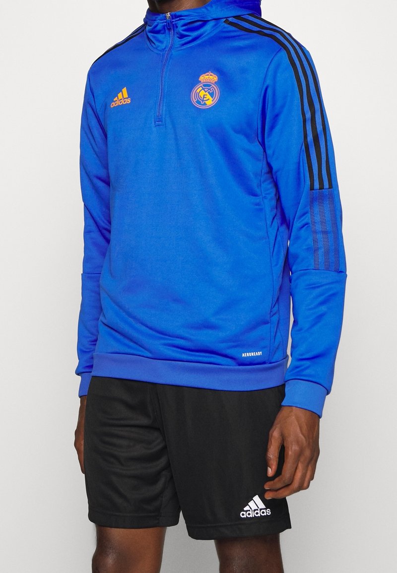 Homme portant un haut d'entraînement bleu Adidas du Real Madrid avec un short noir Adidas sur fond blanc.