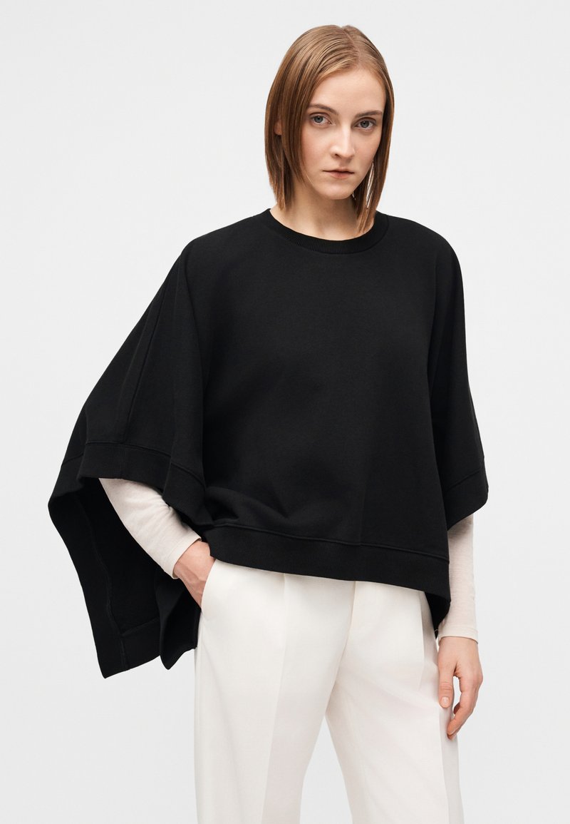 Sweat-shirt noir surdimensionné avec manches larges, encolure côtelée et tissu texturé, superposé à un haut à manches longues de couleur claire.