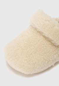 Fuzzy krämfärgad toffel med mjuk textur, rund form och vikbar manschett. Lämplig för inomhusbruk.