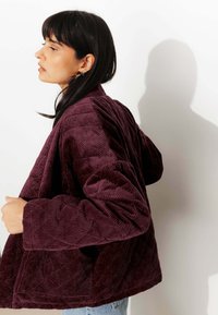 Veste en velours côtelé bordeaux avec texture matelassée, dotée de larges manches et d'une coupe décontractée. Le tissu présente des motifs de nervures distincts.