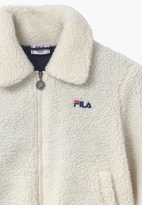 Kremfarvet fleecejakke med krave, lynlås foran og broderet Fila-logo i marineblå og rød på brystet. Blød tekstur.