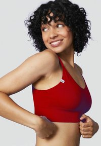 Reggiseno sportivo rosso a coste con spalline regolabili, caratterizzato da un piccolo etichetta su un lato. Indossato da un modello con capelli ricci. Texture liscia.