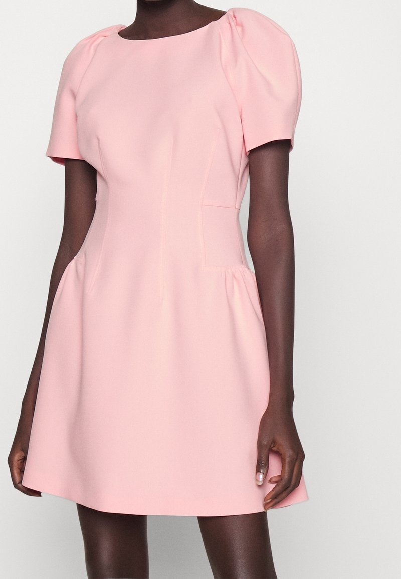 Femme portant une robe courte rose clair avec des manches courtes bouffantes et une taille cintrée, debout devant un fond uni.