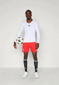 Conjunto desportivo Adidas com uma parte de cima justa de mangas longas brancas, calções vermelhos com o logo do Bayern de Munique e meias pretas com riscas brancas.