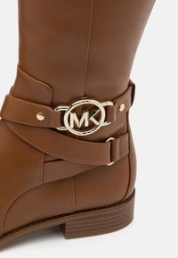 Bottine en cuir marron à bout rond, dotée d'une boucle logo circulaire dorée et de détails de sangle décoratifs. Talon bas empilé.