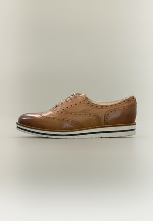 Chaussure de ville brogue en cuir marron, ornée de perforations décoratives, à lacets et dotée d’une semelle blanche à plusieurs couches, présentée de profil sur un fond uni.