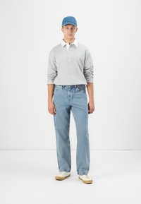 Jauns vīrietis, kurš valkā zilu cepuri, gaiši pelēku garām piedurknēm polo krekli, zilos džinsus un baltas sporta apavus, stāv pret balto fona.