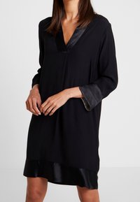 Svart klänning med V-ringning, lång ärm och satin detaljer vid ärmsluten och fållen. Tillverkad av ett mjukt material med en avslappnad passform.
