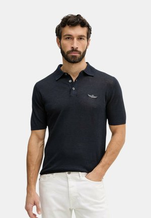 Uomo con la barba che indossa una polo blu scuro a maniche corte con bottoni e pantaloni bianchi, in piedi con una mano in tasca.