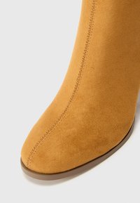 Beige Wildleder-Ankle-Boot mit spitzem Zehenbereich, glatter Textur und sichtbaren Nähten entlang der Nähte. Leicht geschwungener Absatz.